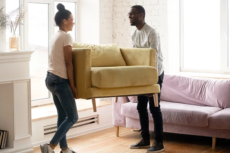 A smiling couple carrying a yellow armchair across a room with old wood floors to prepare for renovation.