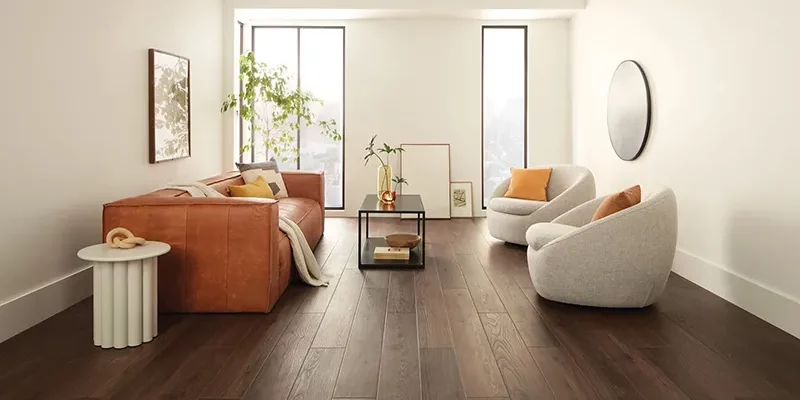A bright, contemporary living room with dark brown hardwood flooring, a cognac leather sofa, and two curved off-white accent chairs.