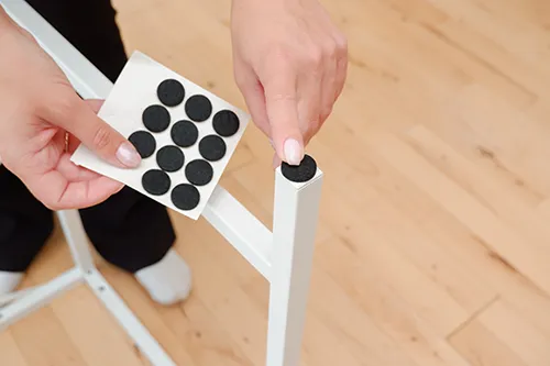 Close-up of a person applying a circular black felt pad to the bottom of a white furniture leg to protect the floor.