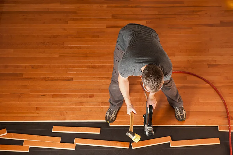 Overhead view of an installer using a pneumatic flooring nailer and a mallet to lay down warm-toned hardwood planks.