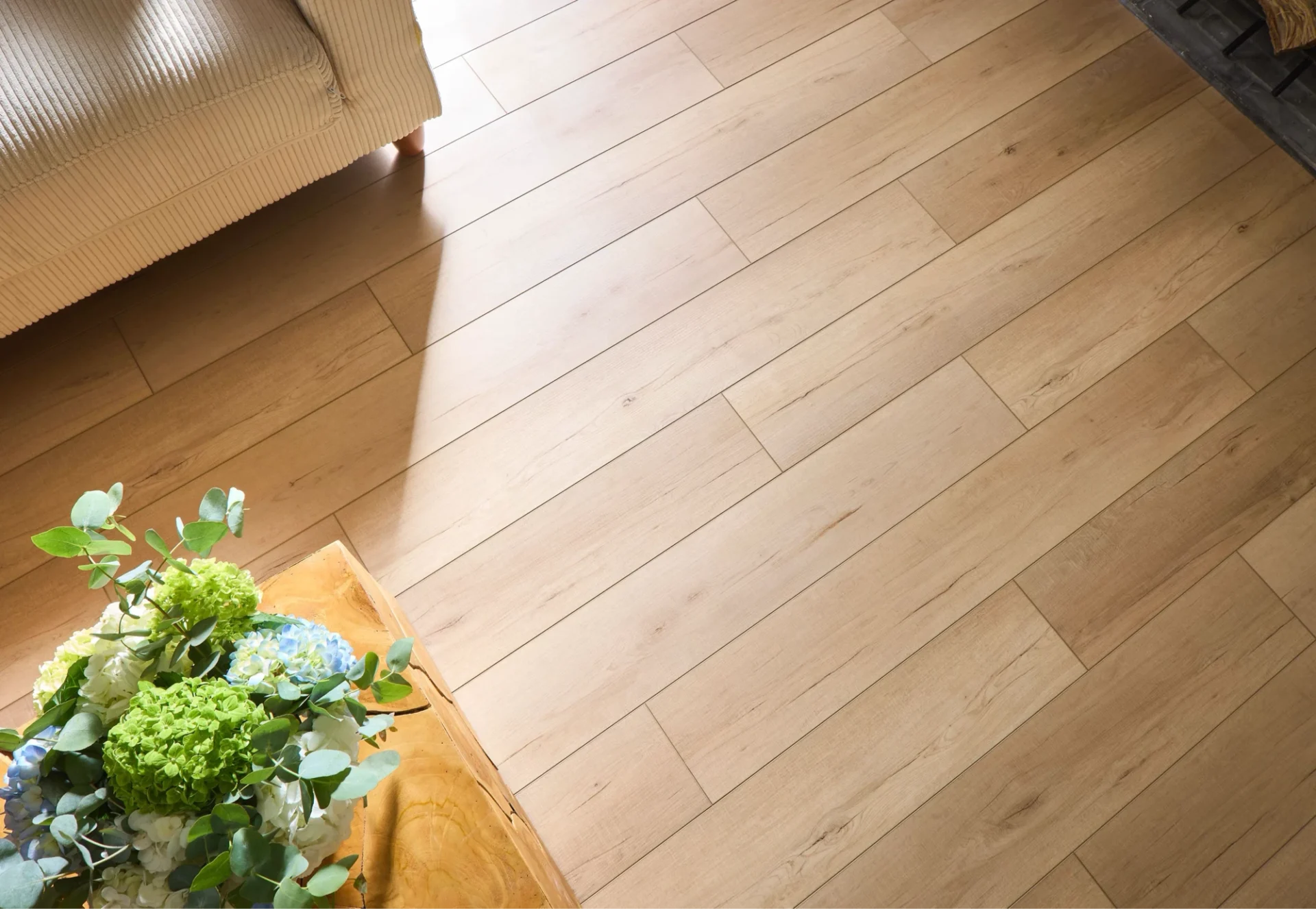 A high-angle view of light oak-look flooring planks, with the corner of a cream corduroy sofa and a wooden table holding a vibrant green and blue floral arrangement.