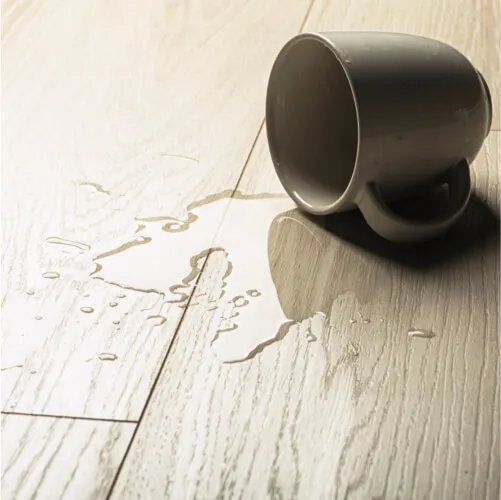 Close-up of a tipped-over ceramic mug spilling clear liquid onto a light natural wood floor.