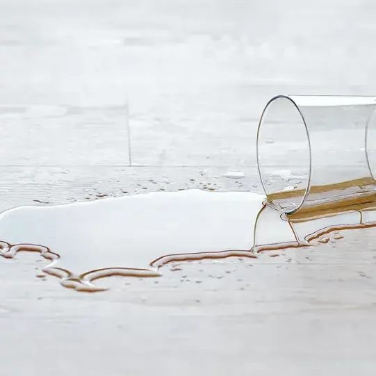 A puddle of dark amber liquid spilled onto a light grey wood-look laminate floor from an overturned glass.