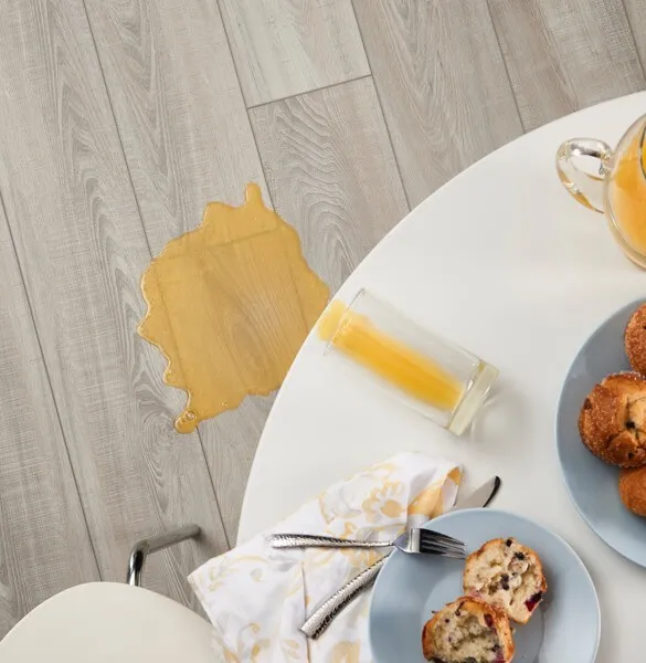 Overhead view of orange juice spilled from an overturned glass onto light grey wood-look flooring next to a round white dining table.